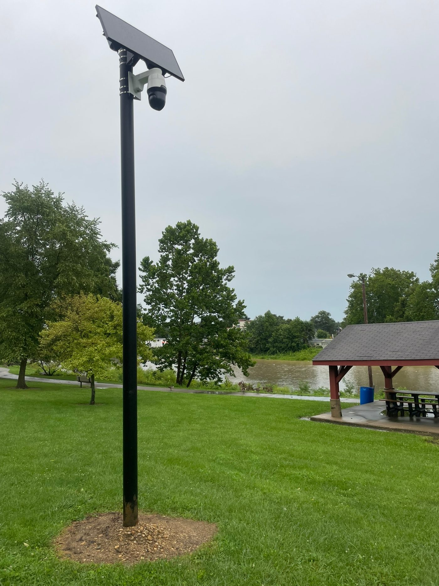 The camera on North River Drive in Marion, photographed by OurWeek at 3:28 p.m. on August 11, 2026. The paired Live Photo files place the shot at 40.5738, -85.6618 — five feet from the camera's mapped location. A white unit with a black head, on its own solar mast in the park lawn: the asphalt path crosses the frame and a public picnic shelter stands at right, with the Mississinewa behind.